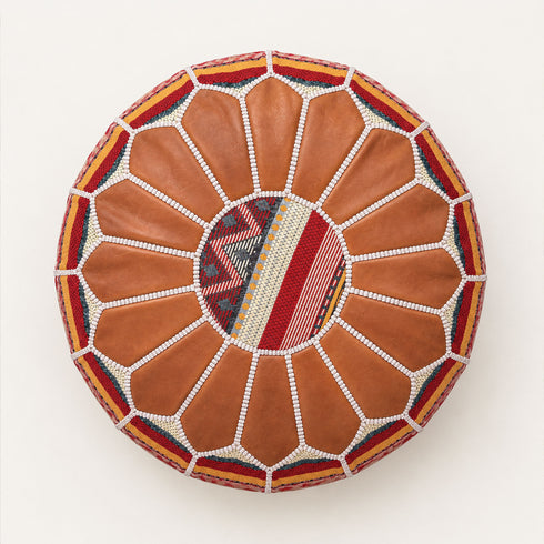Top view of a stuffed tan round fusion pouf with leather panels, white beaded stitching and a center of red, gold and cream Amazigh Berber motif fabric