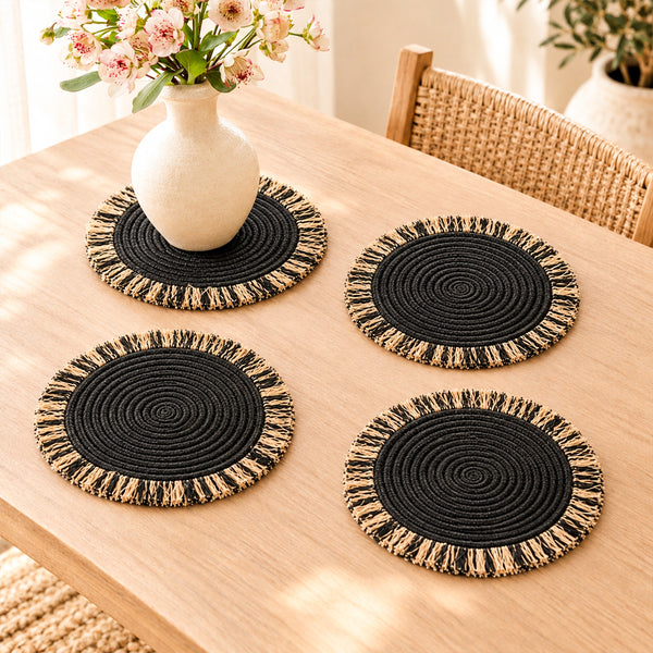 Four black woven table mats with natural and black raffia fringe on an oak table, one under a flower vase
