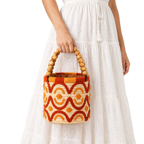 Woman holding handmade Moroccan raffia bucket bag by its wooden bead top handle in a white outfit
