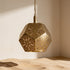 A brass geometric pendant light with intricate laser-cut floral patterns, hanging over a sandy surface in a warm, minimalist room corner.
