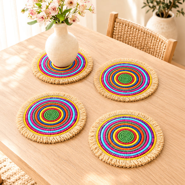 Four colorful concentric woven placemats with natural fringe on an oak table, one used under a flower vase