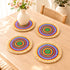 Four colorful concentric woven placemats with natural fringe on an oak table, one used under a flower vase