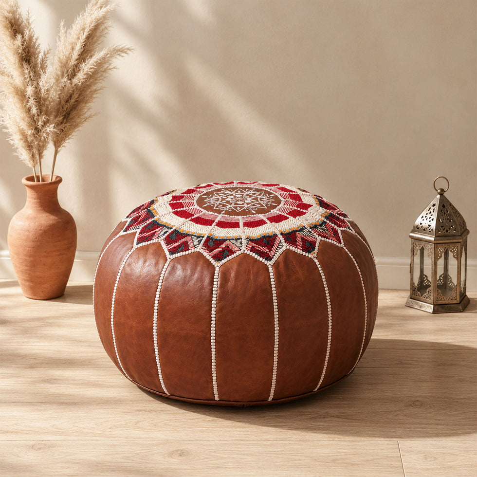 Dark tan round fusion Moroccan leather pouf with red Amazigh Berber motif fabric top beside pampas grass and a Moroccan lantern