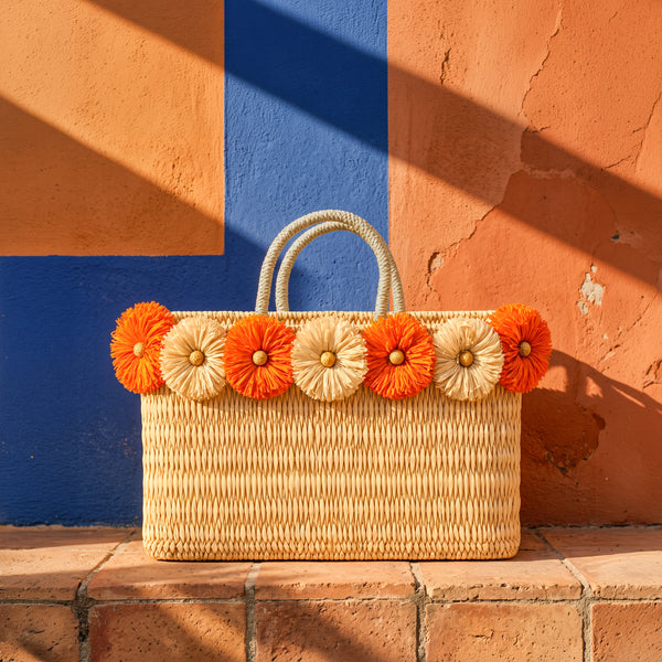 Orange raffia flower wicker tote bag with braided handles on a terracotta tile ledge against a cobalt blue Moroccan wall