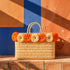 Orange raffia flower wicker tote bag with braided handles on a terracotta tile ledge against a cobalt blue Moroccan wall