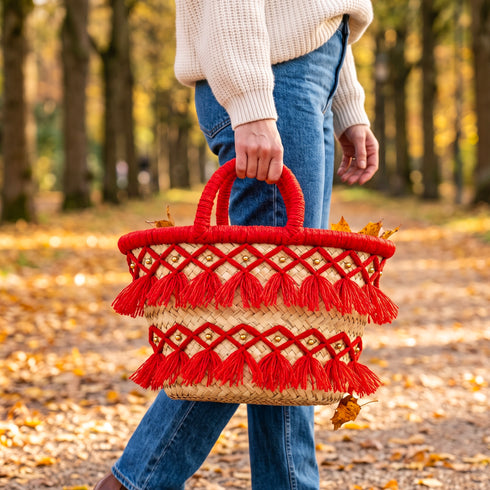 Model carrying a handmade fire red tassel straw basket with gold beads on an autumn forest path with fallen leaves