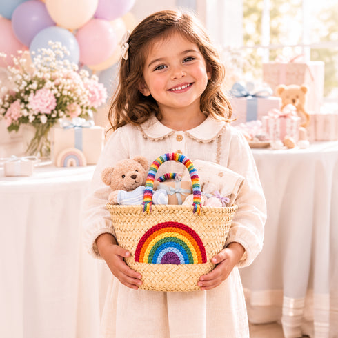 Smiling little girl holding a rainbow baby shower basket with embroidered rainbow, teddy bear and party gifts