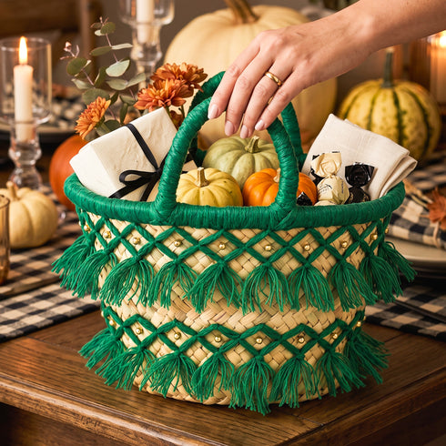 Deep green tassel straw basket filled with mini pumpkins, napkins and a wrapped gift as a Thanksgiving table centerpiece