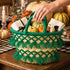 Deep green tassel straw basket filled with mini pumpkins, napkins and a wrapped gift as a Thanksgiving table centerpiece