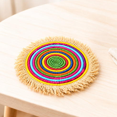 Handwoven Moroccan rainbow raffia Small placemat with colorful rings and natural fringe on a pale wooden table