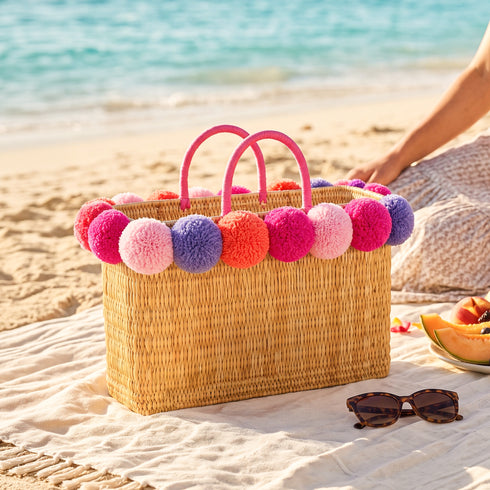 Multicolor pom pom wicker beach bag in pink, lilac and fuchsia on a beach towel with sunglasses, a bachelorette party bag