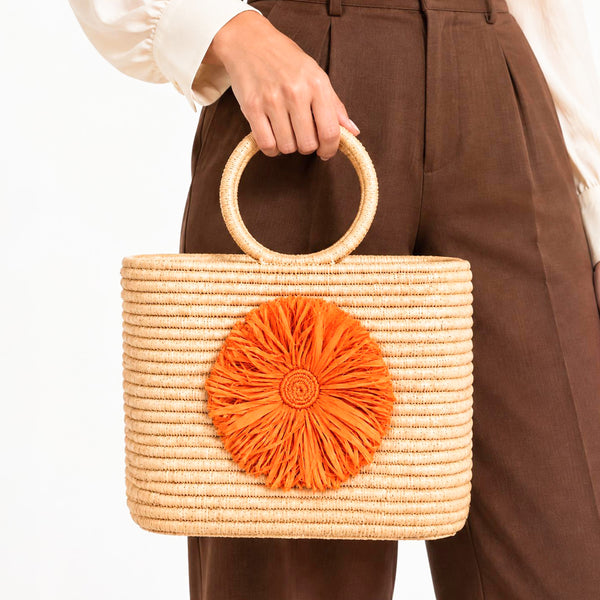 Natural round handle raffia tote bag with orange raffia flower held with brown trousers and a cream blouse for fall