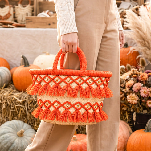 Model holding an orange Moroccan tassel straw market basket with gold beads beside pumpkins, hay bales and dried flowers