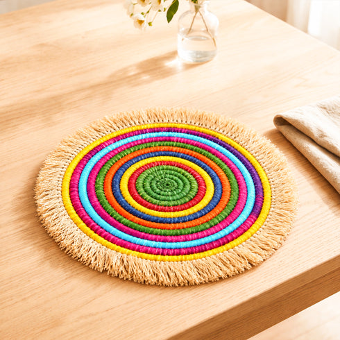 Rainbow concentric woven raffia Large placemat with natural fringe on a light oak table beside a linen napkin