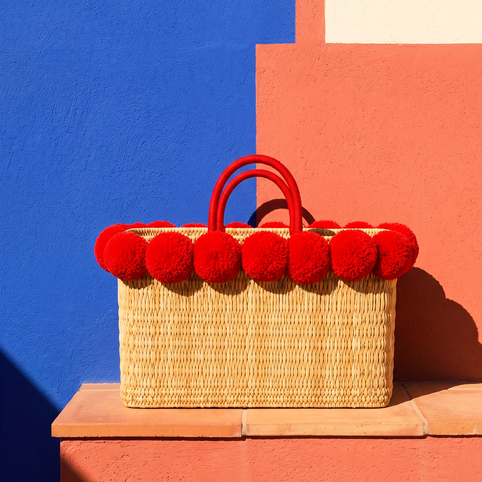 Red pom pom straw basket bag with matching double handles against a cobalt blue and coral Moroccan wall