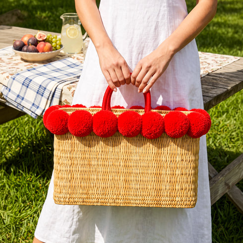 Woman in white linen dress holding a red pom pom wicker bag by its matching double handles at a garden picnic