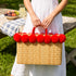Woman in white linen dress holding a red pom pom wicker bag by its matching double handles at a garden picnic
