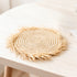 Round woven placemat with uneven raffia fringe edge and beige Doum palm spiral weave on a pale wooden table