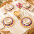 Set of 4 rainbow woven raffia placemats with cream plates and bowls on a linen-covered table with pink flowers