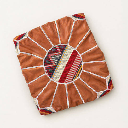Unstuffed tan round fusion pouf cover laid flat, showing leather panels with a center of red, cream and gold Moroccan Amazigh Berber motif fabric