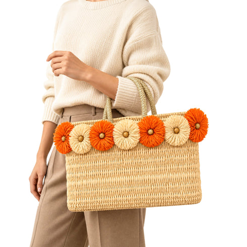 Woman in cream sweater and beige trousers carrying a wicker tote bag with orange and natural raffia flowers on the rim