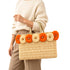 Woman in cream sweater and beige trousers carrying a wicker tote bag with orange and natural raffia flowers on the rim