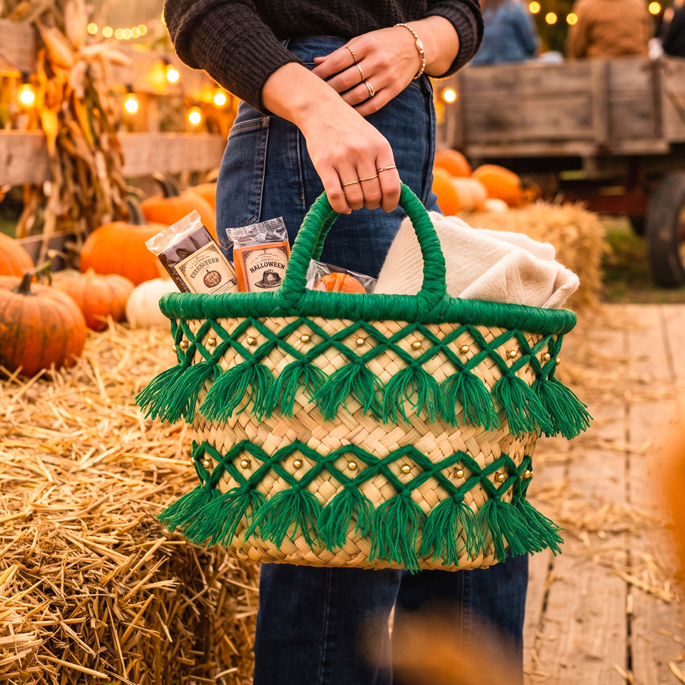 Model holding a handmade green tassel straw basket with treats and a cream blanket beside pumpkins and hay bales at a fall farm